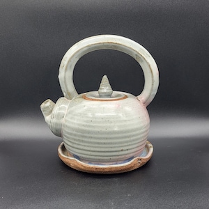 May include: A small, gray ceramic teapot with a pink lid and a matching saucer. The teapot has a rounded body and a handle that curves up and over the top.