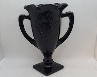 Peut inclure: Un vase en verre noir avec deux anses et un motif orné. Le vase a une base carrée et un sommet cannelé.