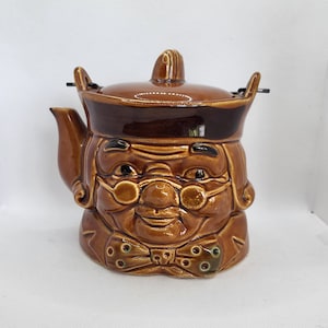 Puede incluir: Una tetera de cerámica marrón con forma de cabeza de persona con un sombrero marrón, gafas y una pajarita. La tetera tiene un pico y una tapa.