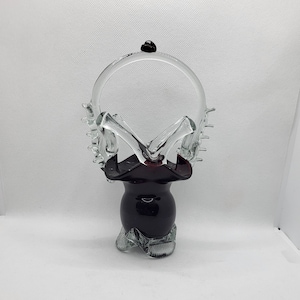 Könnte beinhalten: Ein klarer Glaskorb mit dunkelrotem Boden und Griff. Der Griff hat eine kreisförmige Form mit einem kleinen schwarzen Akzent oben. Der Korb hat ein einzigartiges, dekoratives Design, perfekt zum Ausstellen kleiner Gegenstände oder als dekoratives Stück.