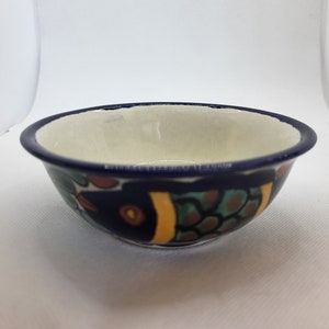 Puede incluir: Un pequeño cuenco de cerámica blanca con un borde azul y un diseño de pez colorido. El cuenco está decorado con pintura azul, verde, amarilla y marrón.