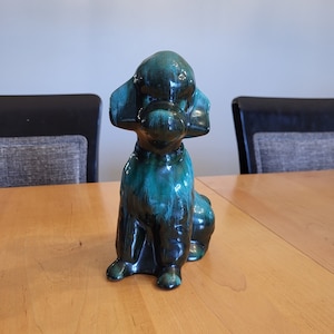 May include: A green glazed ceramic poodle figurine. The poodle is sitting with its head tilted slightly to the side. The glaze has a mottled effect, with shades of dark green and light green.