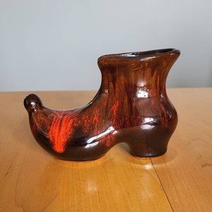 Peut inclure: Un vase en céramique en forme de botte avec un glaçage brun et orange. Le vase a une pointe pointue et un haut évasé.