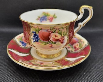 May include: A vintage teacup and saucer set with a floral design featuring peaches, grapes, and berries. The cup and saucer are decorated with gold trim and a red and pink background.