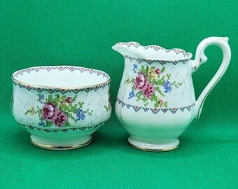 Può includere: Un set di zuccheriera e lattiera bianca con un motivo floreale. Il motivo presenta fiori rosa, rossi e blu su uno sfondo bianco.