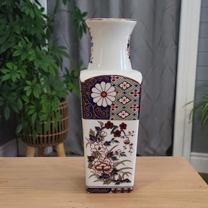 May include: A white ceramic vase with a geometric and floral design. The vase has a square base and a narrow neck. The design features a large white flower with a yellow centre, surrounded by smaller flowers and leaves in shades of red, blue, and green.