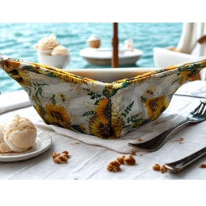 May include: A white ceramic bowl with a floral bowl cosy featuring sunflowers and bees. The bowl is on a white tablecloth with a fork and a plate of ice cream. The background shows the ocean and other dishes.