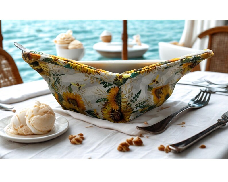 May include: A white ceramic bowl with a floral bowl cosy featuring sunflowers and bees. The bowl is on a white tablecloth with a fork and a plate of ice cream. The background shows the ocean and other dishes.