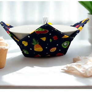 May include: A white ceramic bowl is nestled in a navy blue bowl cosy. The cosy features colourful fruit illustrations, such as pineapples, kiwi, and watermelon. A spoon rests in the bowl. A scoop of ice cream is to the left.
