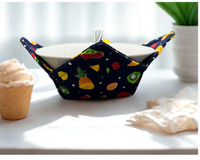 May include: A white ceramic bowl is nestled in a navy blue bowl cosy. The cosy features colourful fruit illustrations, such as pineapples, kiwi, and watermelon. A spoon rests in the bowl. A scoop of ice cream is to the left.