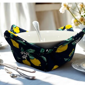 May include: A white ceramic bowl with a silver spoon inside, set in a navy blue bowl cosy. The cosy features a pattern of yellow lemons, green leaves, and white flowers. The scene is on a white tablecloth with silverware.