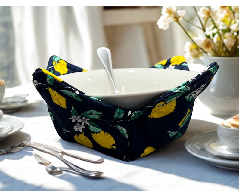May include: A white ceramic bowl with a silver spoon inside, set in a navy blue bowl cosy. The cosy features a pattern of yellow lemons, green leaves, and white flowers. The scene is on a white tablecloth with silverware.