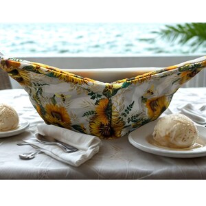 May include: A white bowl cosy with a sunflower pattern, holding a white bowl. The cosy has yellow, white, and green floral designs. Two plates of ice cream and silverware are on the table.