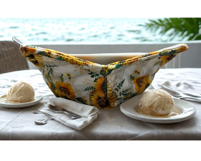 May include: A white bowl cosy with a sunflower pattern, holding a white bowl. The cosy has yellow, white, and green floral designs. Two plates of ice cream and silverware are on the table.