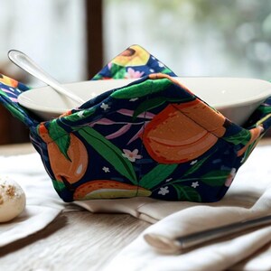 May include: A navy blue bowl cosy featuring a colourful orange and green fruit design. It holds a white bowl with a spoon. The cosy sits on a white napkin, with a fork and pastries in the background.