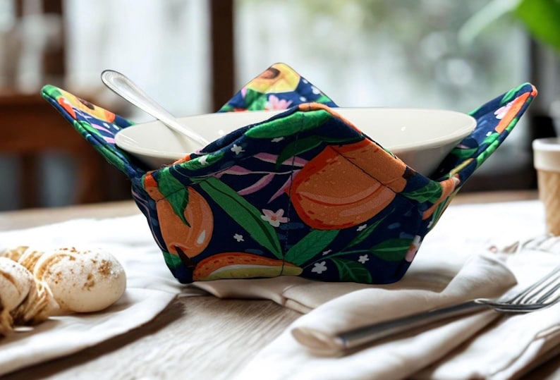 May include: A navy blue bowl cosy featuring a colourful orange and green fruit design. It holds a white bowl with a spoon. The cosy sits on a white napkin, with a fork and pastries in the background.