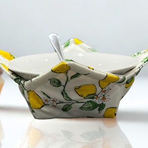 May include: A white ceramic bowl with a spoon sits within a quilted fabric bowl cosy. The cosy features a lemon and floral pattern with yellow lemons, green leaves, and white blossoms on a light grey background. A cupcake is in the background.