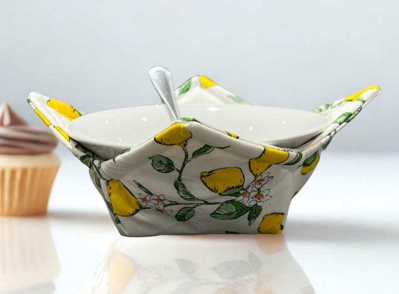 May include: A white ceramic bowl with a spoon sits within a quilted fabric bowl cosy. The cosy features a lemon and floral pattern with yellow lemons, green leaves, and white blossoms on a light grey background. A cupcake is in the background.