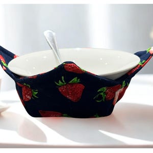 May include: A navy blue bowl cosy featuring a red strawberry design, holding a white ceramic bowl and a silver spoon. The cosy is designed to keep bowls warm and is placed on a white surface.
