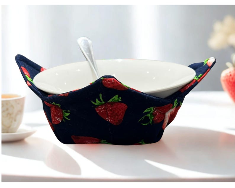 May include: A navy blue bowl cosy featuring a red strawberry design, holding a white ceramic bowl and a silver spoon. The cosy is designed to keep bowls warm and is placed on a white surface.