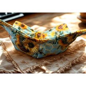 May include: A fabric bowl cosy with a sunflower pattern. The cosy features a light blue background with yellow sunflowers and a honeycomb design. The edges are folded to create a bowl shape, with the sunflower pattern visible on the inside and outside.