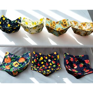 May include: A selection of fabric bowl cozies featuring fruit and floral designs. Patterns include lemons, sunflowers, peaches, strawberries, and mixed fruits on a dark backdrop. The cozies are displayed on a white surface.