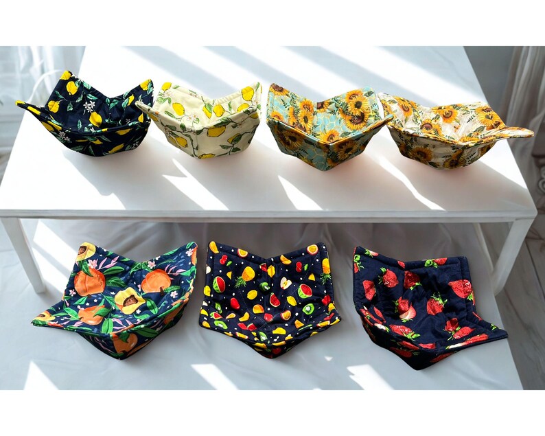 May include: A selection of fabric bowl cozies featuring fruit and floral designs. Patterns include lemons, sunflowers, peaches, strawberries, and mixed fruits on a dark backdrop. The cozies are displayed on a white surface.
