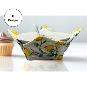 May include: A fabric bowl cosy featuring a white background with a yellow lemon and green leaf pattern, holding a white ceramic bowl with a silver spoon inside. A circular graphic in the upper left corner displays the text 8 Designs.