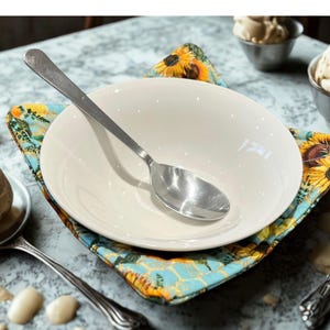 May include: A white ceramic bowl with a stainless steel spoon sits on a colourful, square, floral-patterned placemat. The placemat features sunflowers and a blue background. Ice cream in small metal bowls is visible in the background.