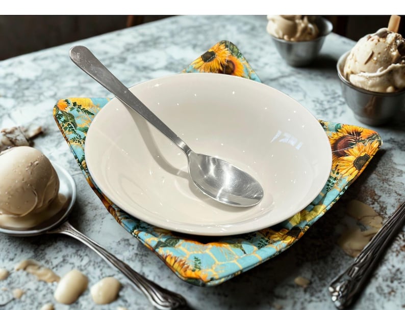 May include: A white ceramic bowl with a stainless steel spoon sits on a colourful, square, floral-patterned placemat. The placemat features sunflowers and a blue background. Ice cream in small metal bowls is visible in the background.