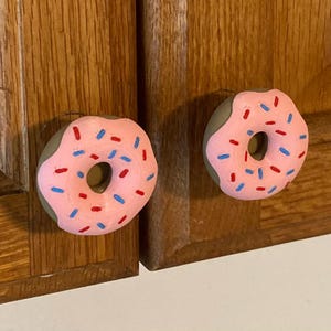 May include: Two donut-shaped cabinet knobs. The knobs have a pink frosting with red and blue sprinkles. The knobs are attached to a wooden cabinet door.