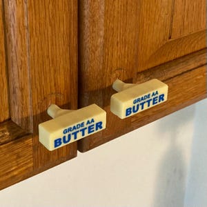 May include: Two rectangular cabinet knobs shaped like sticks of butter. Each knob is off-white with blue text that reads GRADE AA BUTTER. The knobs are attached to wooden cabinet doors, serving as functional handles for opening the doors.