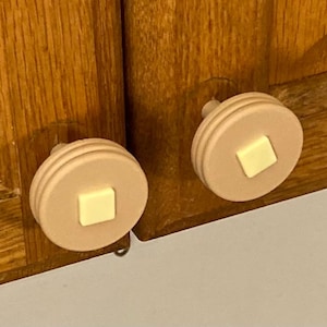 May include: Two cabinet knobs with a pancake design. The knobs are light tan with a square of yellow-beige in the center, resembling a pat of butter. The knobs are attached to a wooden cabinet with a natural finish.