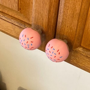 May include: Two round cabinet knobs shaped like pink frosted donuts. Each knob features a smooth, light pink surface decorated with small, scattered red and blue sprinkles. The knobs are mounted onto a wooden cabinet door with a natural wood grain finish.