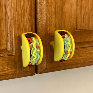 May include: Two cabinet knobs shaped like tacos, featuring a yellow shell, dark brown ground meat, light green lettuce, and small red tomato pieces. Each taco-shaped knob has a wavy yellow line representing cheese or sauce on the front.