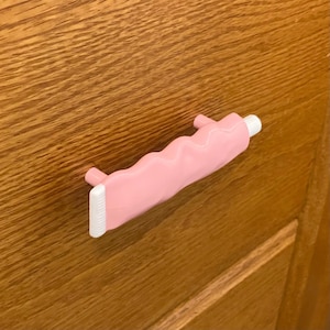 May include: A pink door handle shaped like a tube of toothpaste, with a white cap and a ridged end. The handle is mounted on a wooden door with a visible wood grain.