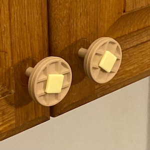 May include: Two wooden cabinet knobs, each shaped like a waffle with a square of yellow butter on top. The knobs are attached to a wooden cabinet with a warm, natural finish. The knobs add a whimsical touch to kitchen decor.