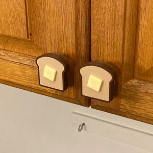 May include: Two cabinet knobs shaped like slices of toast with a square of butter. The knobs are brown, tan, and yellow, and are attached to a wooden cabinet. The knobs are a fun and unique kitchen accessory.