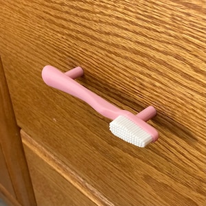 May include: A pink toothbrush-shaped cabinet pull with white bristles. The handle has two cylindrical posts for mounting. The pull is set against a wooden cabinet.