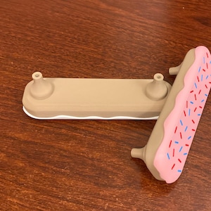 May include: A tan-colored phone stand with two small, rounded posts and a white base. A pink phone grip, resembling a frosted donut with blue and red sprinkles, is also present. The stand and grip are made of a smooth material.
