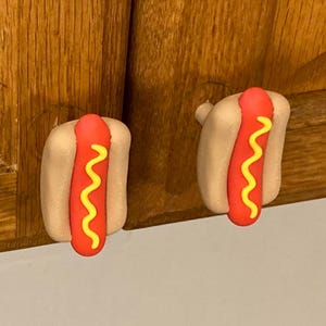 May include: Two hot dog-shaped cabinet knobs. Each knob features a red hot dog with yellow mustard in a beige bun. The knobs are attached to a wooden cabinet.
