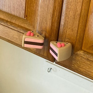 May include: Two cabinet knobs shaped like triangular slices of cake. Each knob features a tan top, a pink middle layer, a dark brown center stripe, and a pink bottom layer. Three small pink dots decorate the top surface of each cake slice knob.