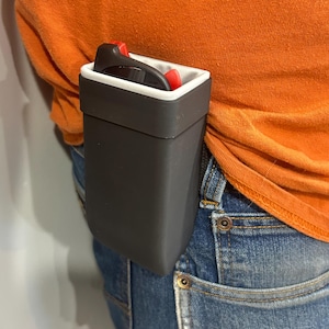 May include: A dark gray sunglasses case with a white interior and red accents is attached to a person's belt. The case is rectangular and appears to be made of plastic. The person is wearing an orange shirt and blue jeans.
