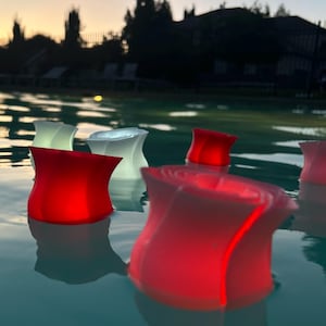 May include: Floating, glowing, LED lights in a pool. The lights are in the shape of a tulip and are red, white, and light green. The water reflects the lights and the sunset in the background.