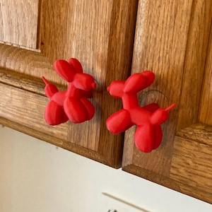 May include: Two bright red cabinet knobs shaped like balloon animals. Each knob features a stylized dog silhouette with rounded ears, a snout, and a tail, designed to function as a handle for furniture doors or drawers with a matte finish.