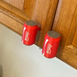 May include: Two red cabinet knobs shaped like miniature Coca-Cola soda cans. Each knob features the white Coca-Cola logo on the side and a grey circular top resembling a can lid. These cylindrical decorative pulls are mounted onto wooden cabinet doors.