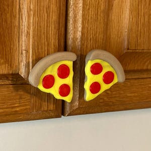 May include: Two cabinet knobs shaped like pizza slices. Each slice has a yellow base with red pepperoni and a brown crust. The knobs are mounted on a wooden cabinet door.