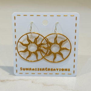 May include: Gold-colored sunburst earrings with silver hooks, displayed on a white card. The earrings feature a circular design with radiating sun rays. The card has the text "SUNRAISERCREATIONS".