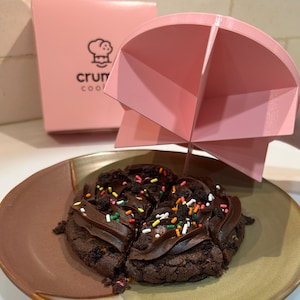 May include: A close-up of a chocolate brownie with colorful sprinkles. The brownie is cut into six pieces and is on a brown plate. A pink plastic dome is above the brownie.