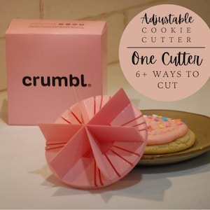 May include: Pink adjustable cookie cutter with the brand name "crumbl." printed on the box. The cutter has six different settings for cutting cookies in various shapes. A pink frosted cookie with sprinkles is shown on a brown plate.